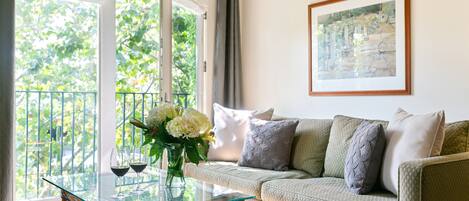 A living room with a glass coffee table, a sofa with cushions, a framed picture on the wall, and a balcony with a view of greenery.