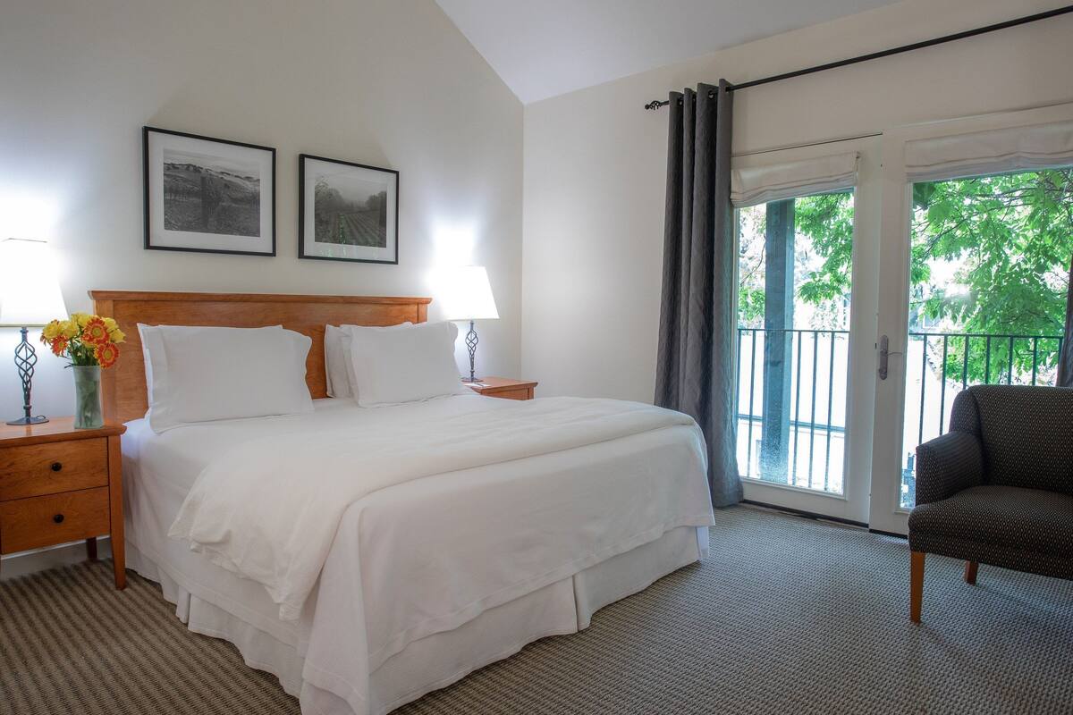 A hotel room with a large bed, a nightstand with a lamp, a chair, and a balcony with curtains.