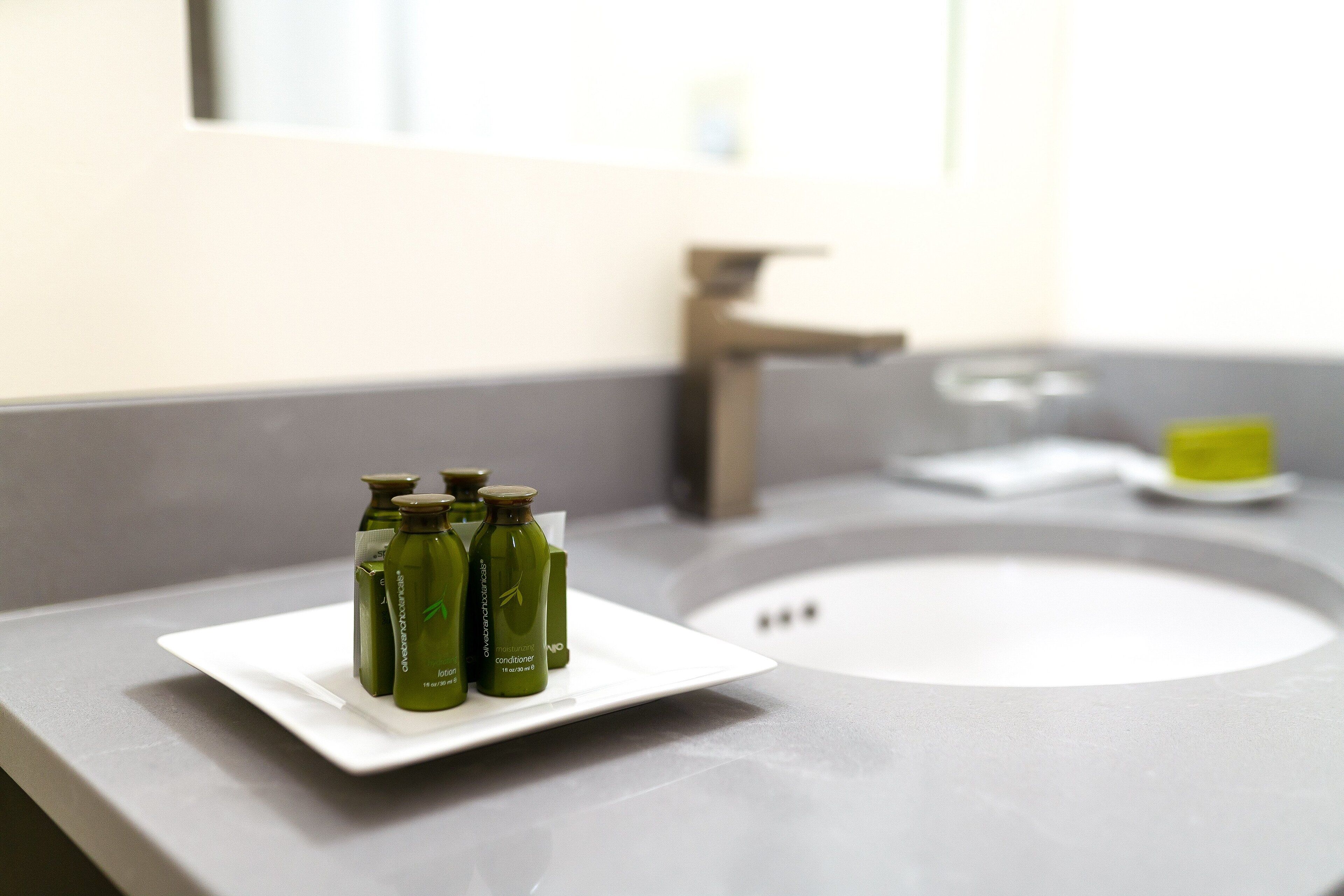 Une salle de bain avec un lavabo blanc, un miroir et trois bouteilles vertes disposées sur un plateau blanc.