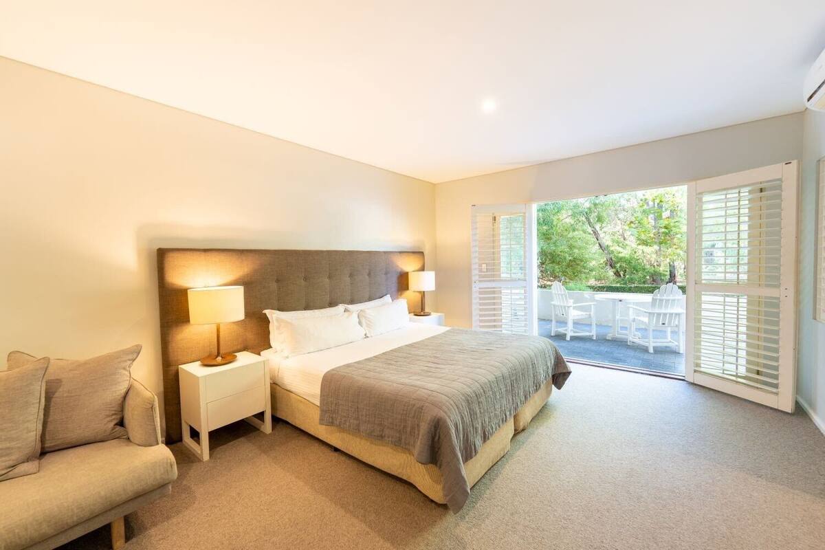 A bedroom with a bed, bedside tables, a chair, and a balcony with outdoor seating.