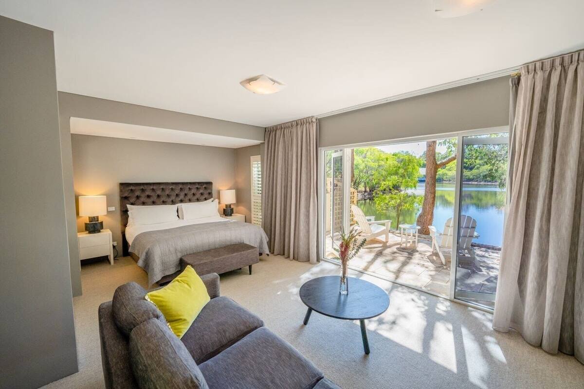 A bedroom with a bed, bedside tables, a sofa, a small table, and a view of a lake through large windows.