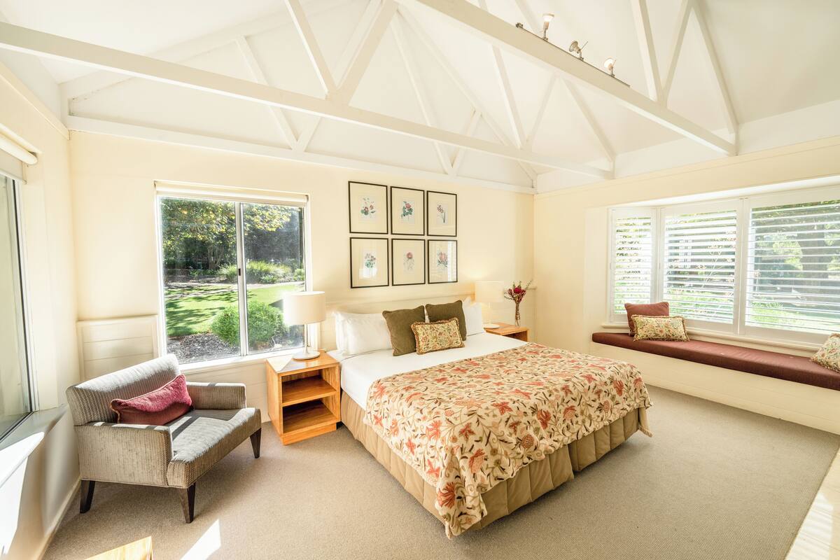 A bedroom with a large bed, a chair, a window with blinds, and a view of greenery outside.