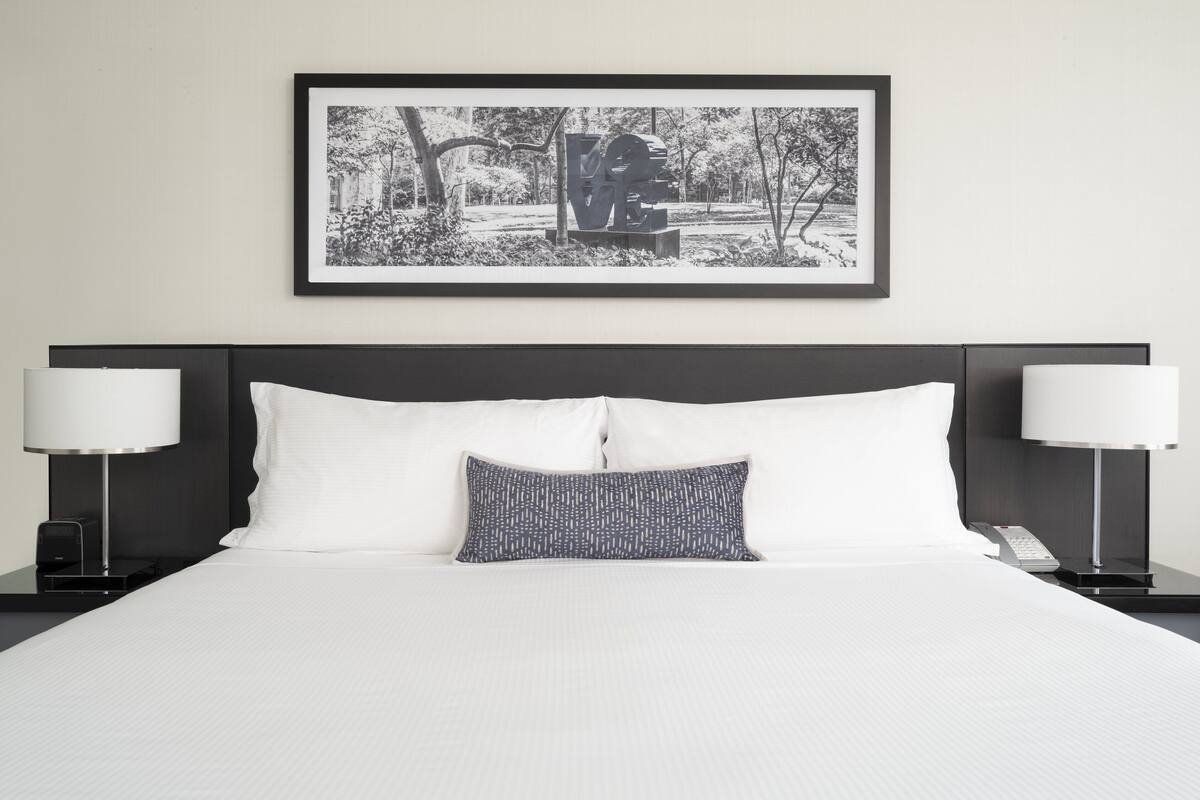 A neatly made bed with white linens and a patterned pillow, flanked by two bedside lamps on black stands. A framed black and white photograph hangs above the bed.