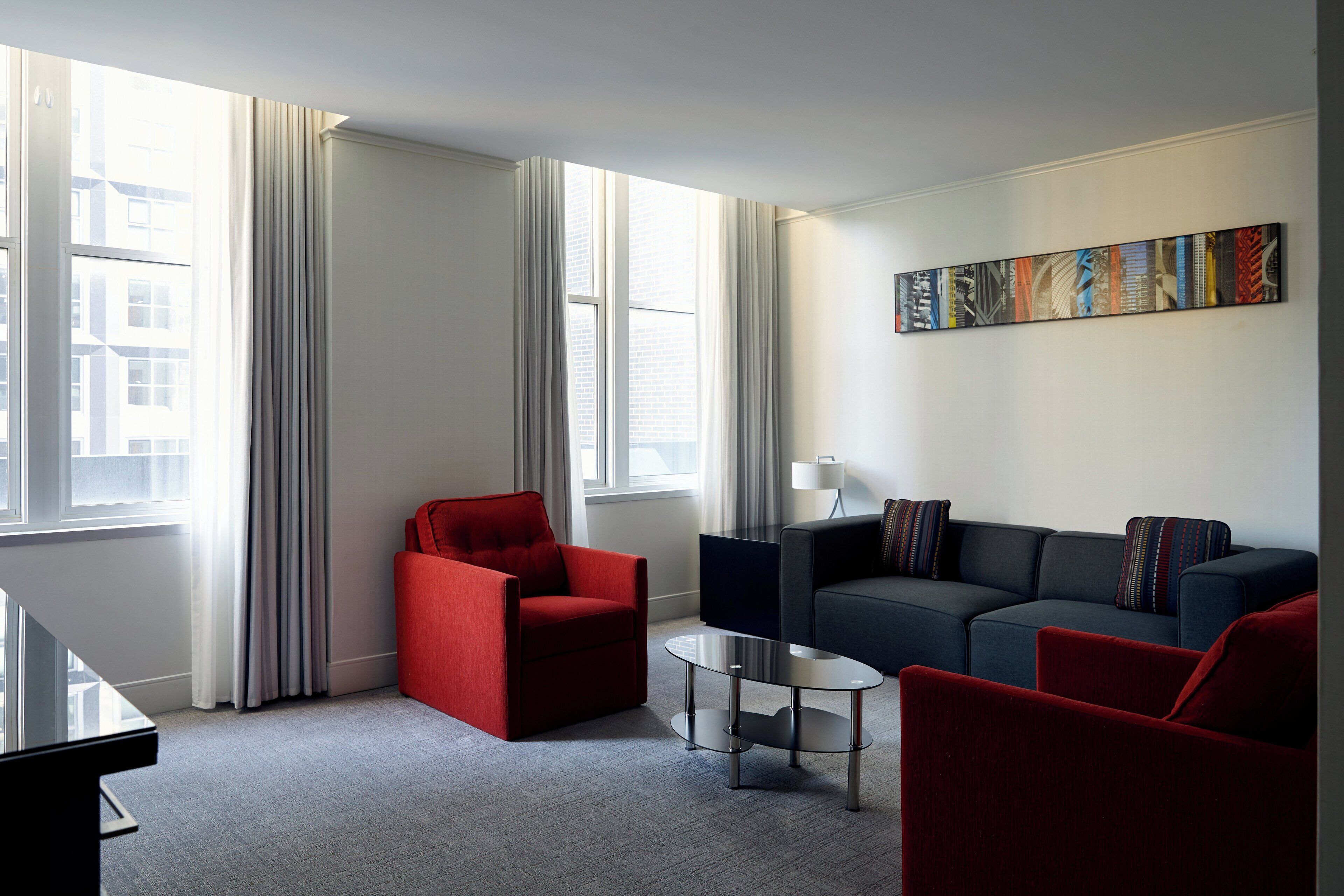 A modern living room with a red armchair, a dark sofa, and a glass coffee table. There is a large window with curtains, a painting on the wall, and a lamp on a side table.