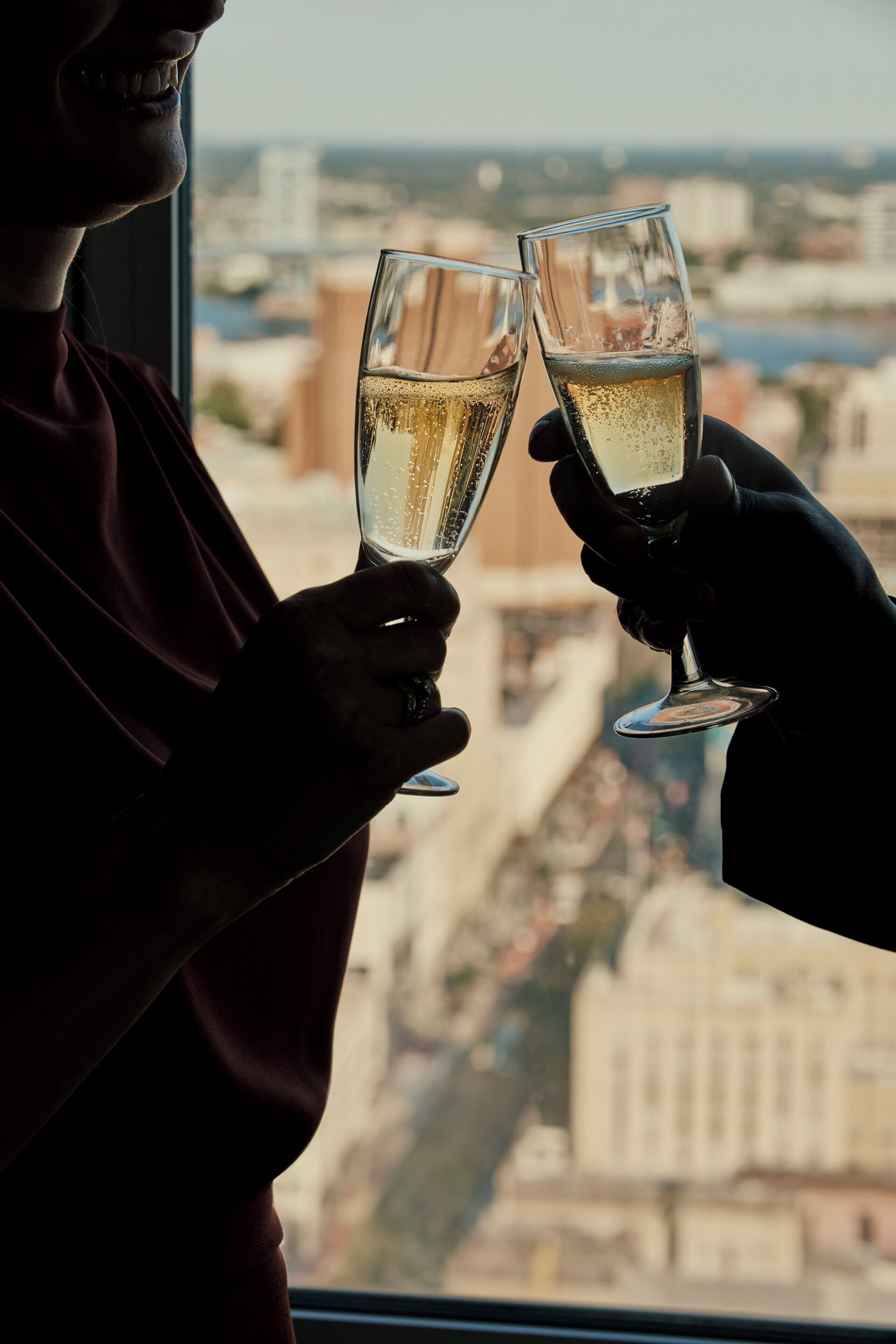 Zwei Personen stoßen mit Champagnergläsern an, im Hintergrund ist eine Stadtlandschaft durch ein Fenster zu sehen.