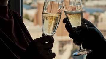 Zwei Personen stoßen mit Champagnergläsern an, im Hintergrund ist eine Stadtlandschaft durch ein Fenster zu sehen.