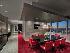 Habitación de hotel moderna con zona de comedor que presenta una mesa grande y sillas rojas, un televisor de pantalla plana en la pared y vistas a la ciudad a través de amplias ventanas.