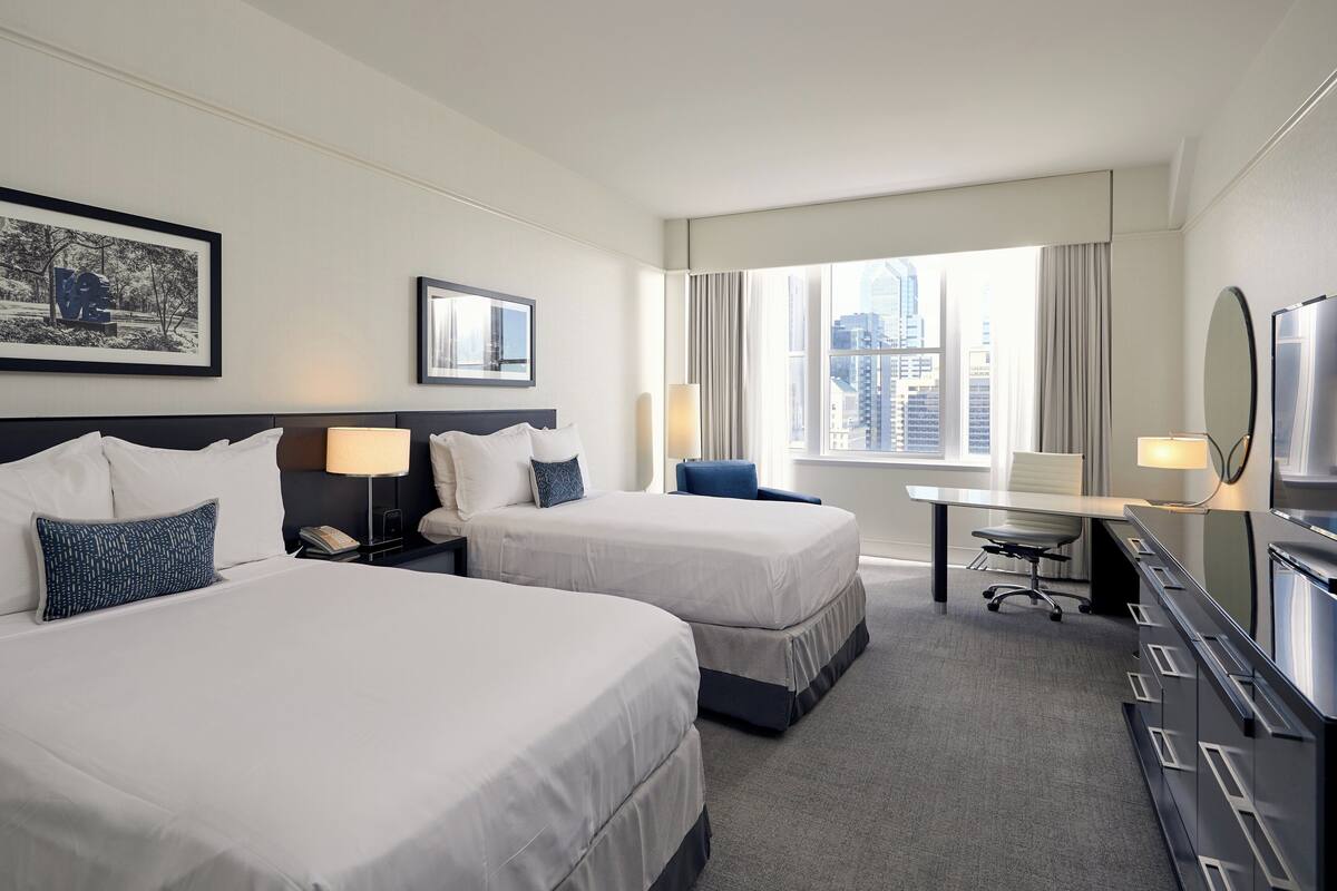 A hotel room with two beds, a desk, a chair, a TV, and a large window with a city view.