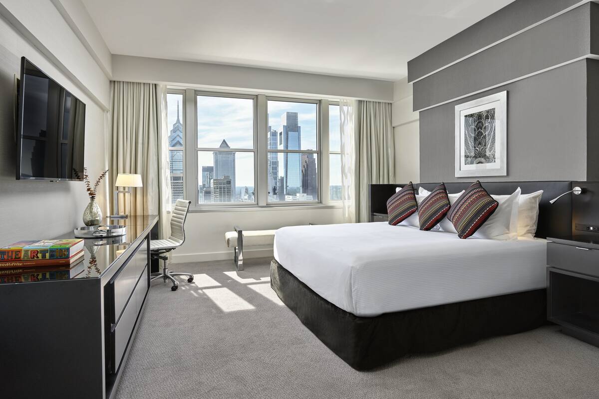 A modern hotel room with a large bed, a desk with a flat-screen TV, a chair, and a view of the city skyline through large windows.