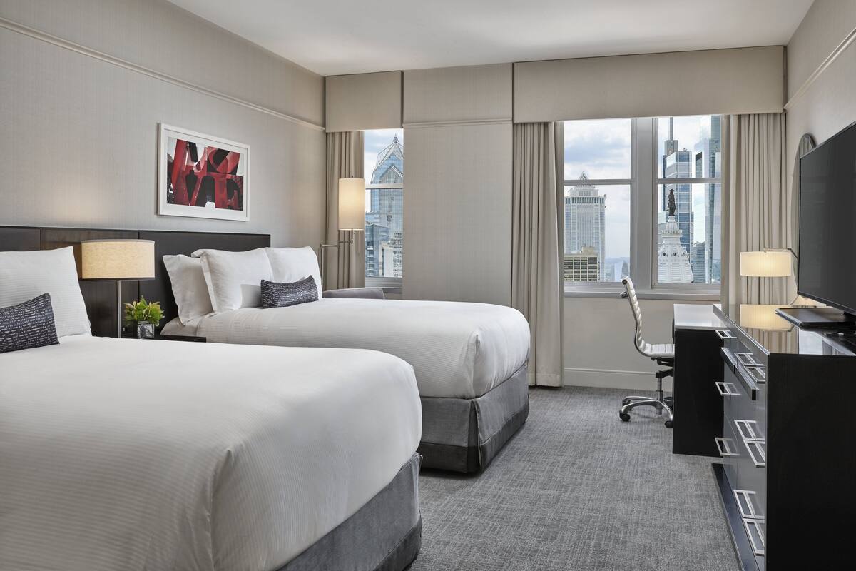 A hotel room with two beds, a desk, a chair, and a large window with a city view.