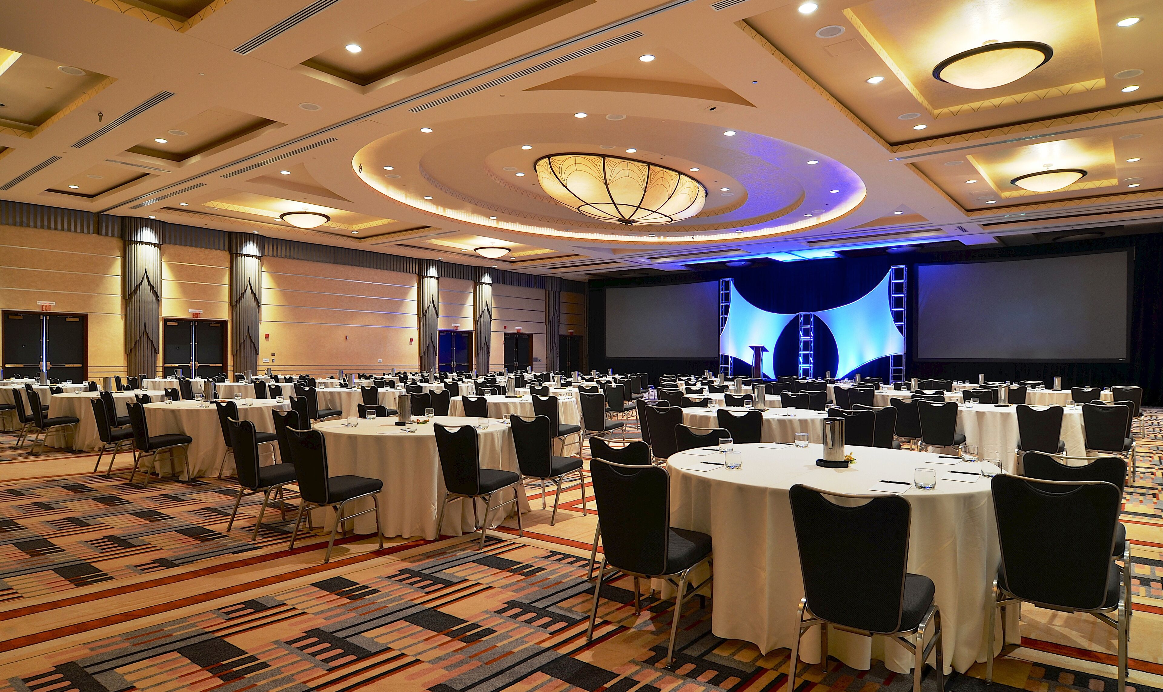 A large conference room with multiple round tables, black chairs, and a stage with a blue screen.