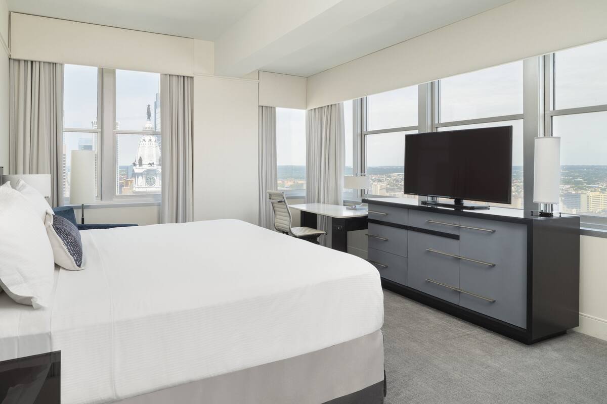 A hotel room with a large bed, a desk, a television, and a view of the cityscape through large windows.