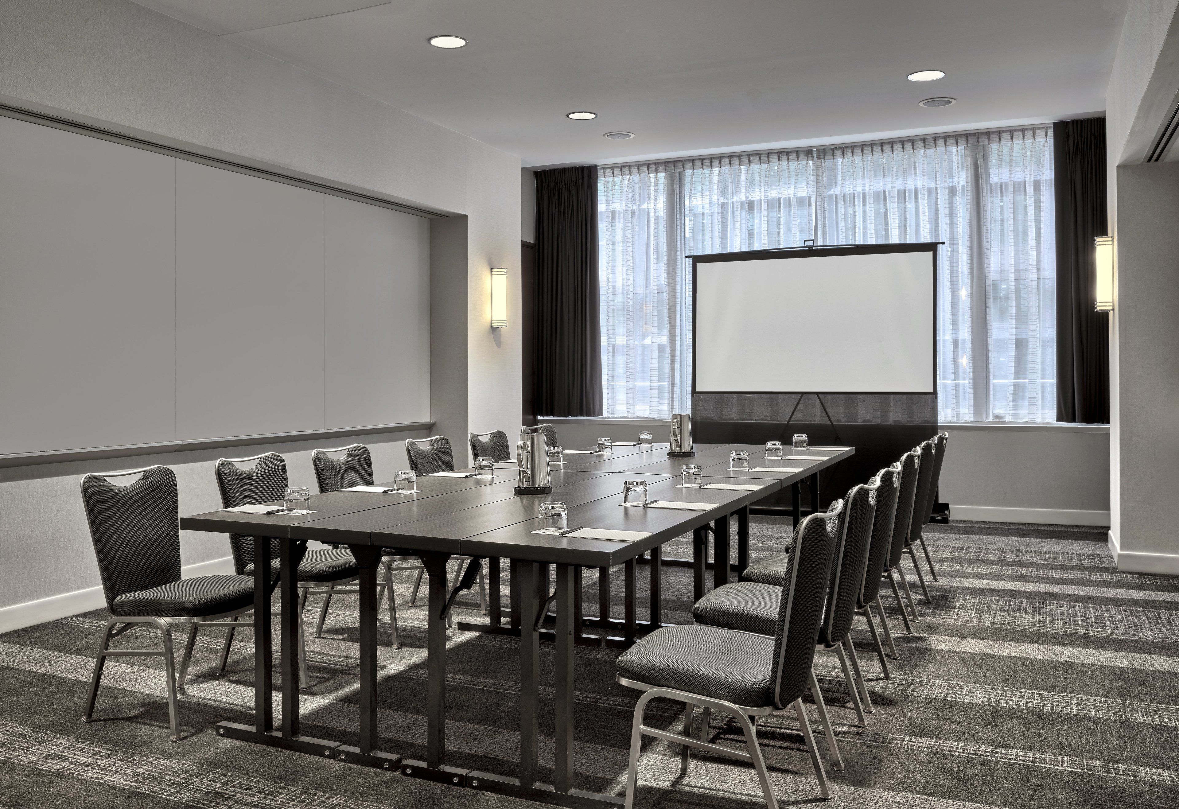 Une salle de conférence avec une longue table, des chaises, un écran de projection et une grande fenêtre avec des rideaux.