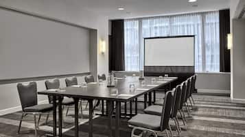 A conference room with a long table, chairs, a projector screen, and a large window with curtains.