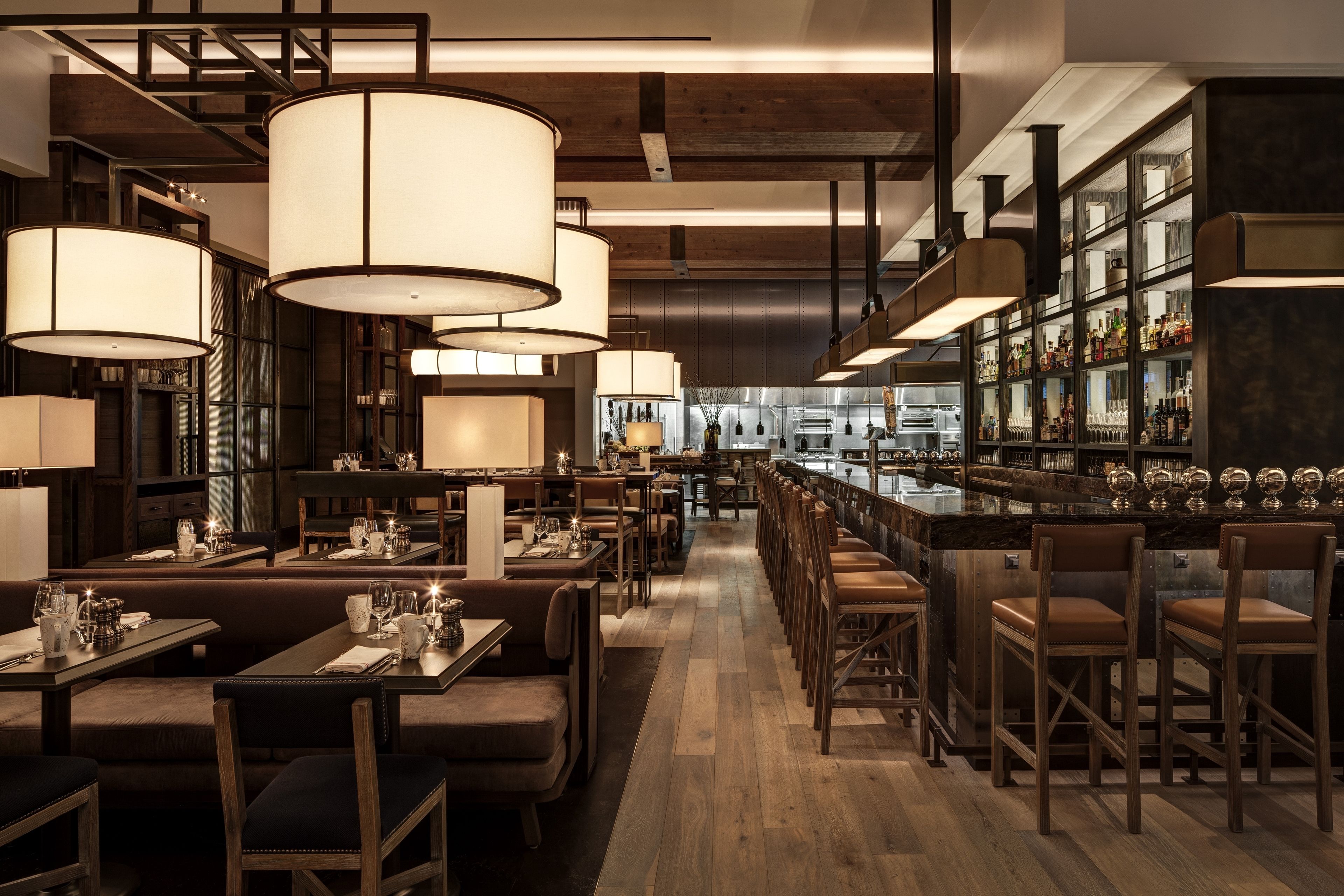 Un restaurante con mesas y sillas de madera, lámparas colgantes grandes y una zona de bar con estantes llenos de botellas.