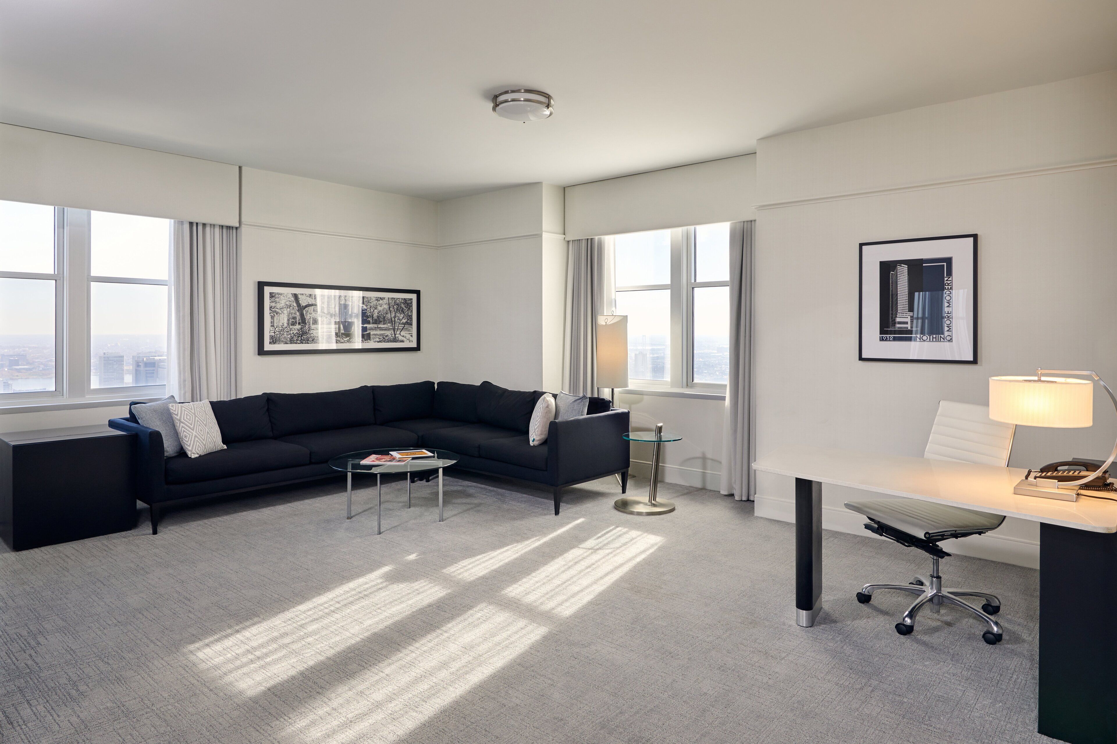 A modern hotel room with a black sectional sofa, a small coffee table, a desk with a lamp, and large windows offering a city view.