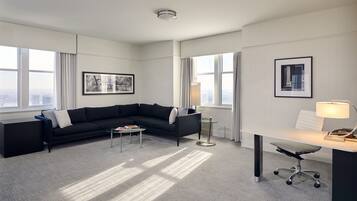 A modern hotel room with a black sectional sofa, a small coffee table, a desk with a lamp, and large windows offering a city view.