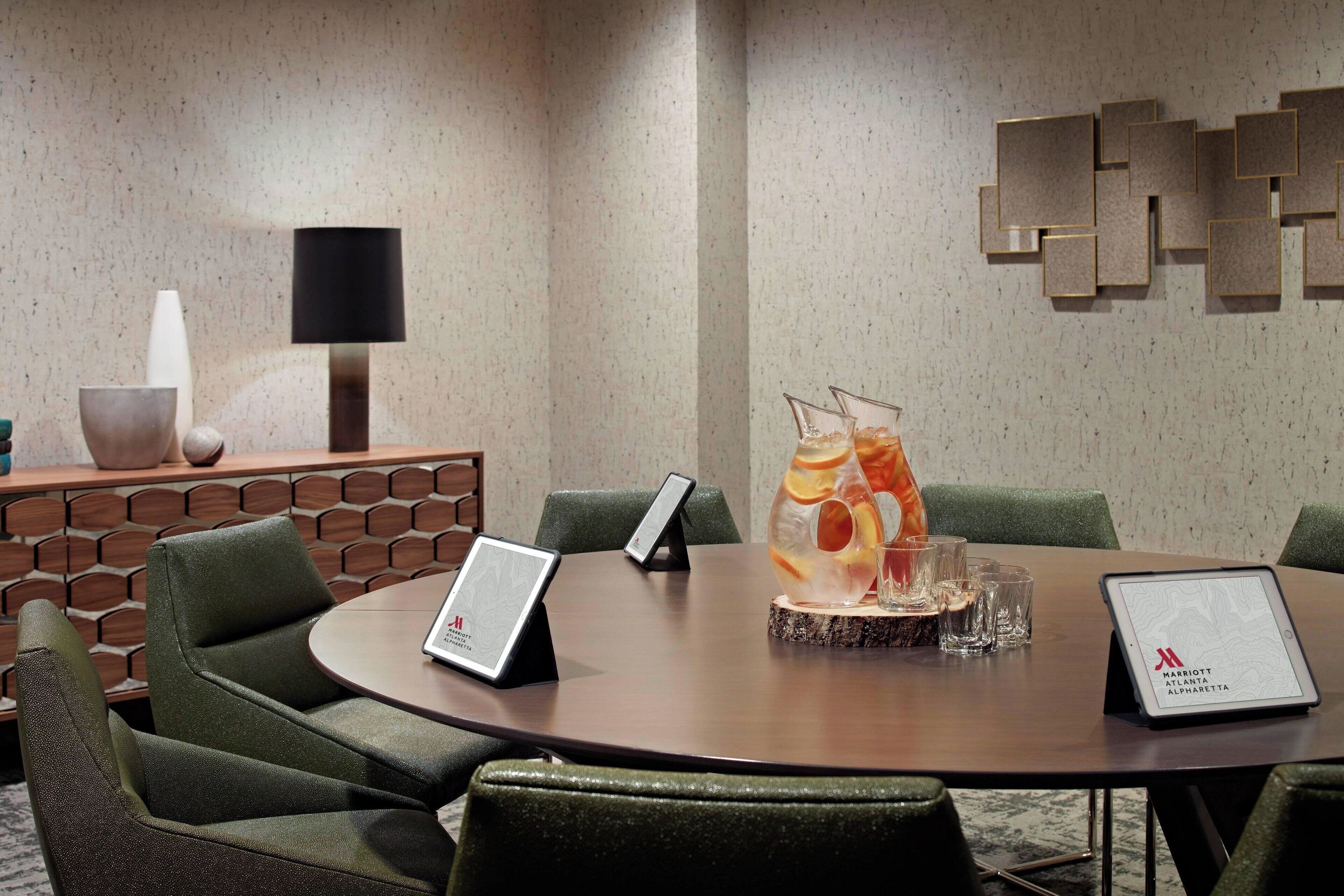 A conference room with a round table, green upholstered chairs, a tablet on the table, and a decorative wall piece.