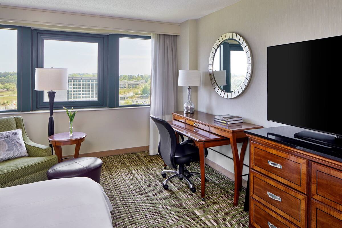 Habitación de hotel con cama, escritorio con computadora, televisor y vista a la ciudad a través de amplias ventanas.