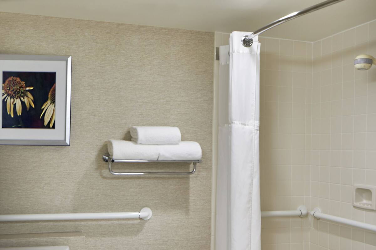 Un baño con inodoro, ducha con cortina blanca, barra de apoyo, pequeño taburete con toalla y estante con toallas.
