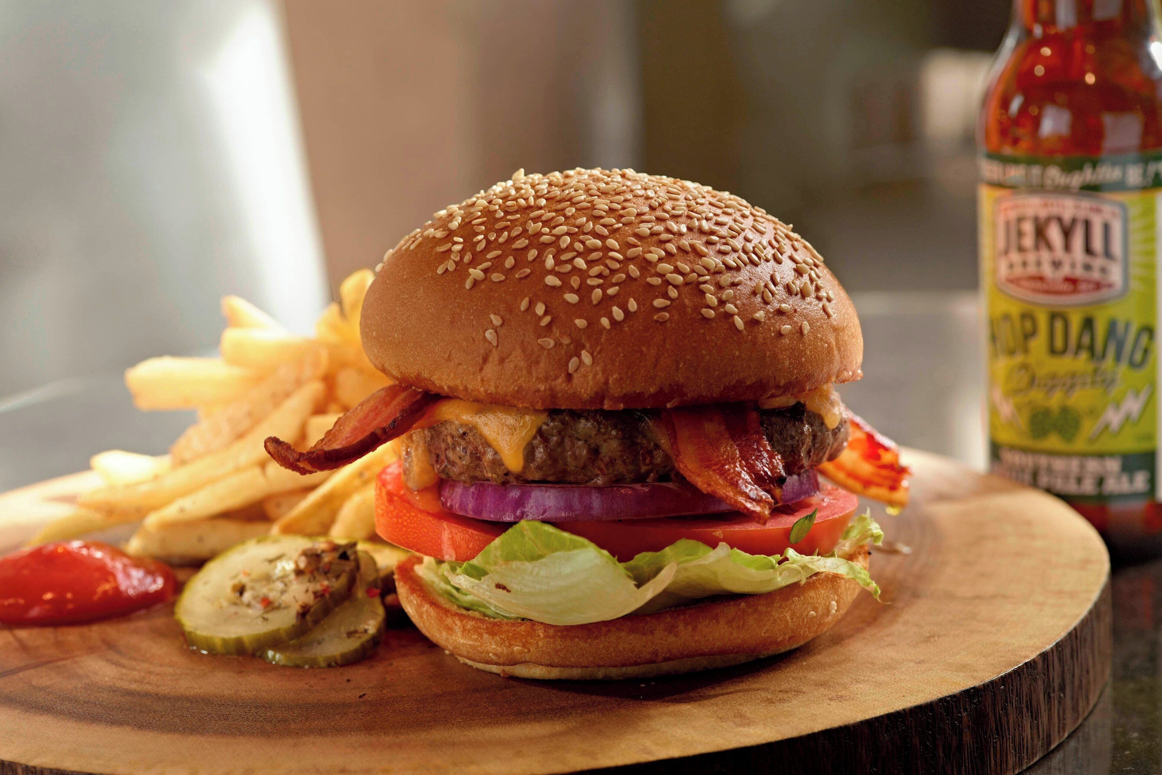 A burger with bacon, cheese, and lettuce on a wooden board, accompanied by fries and pickles.
