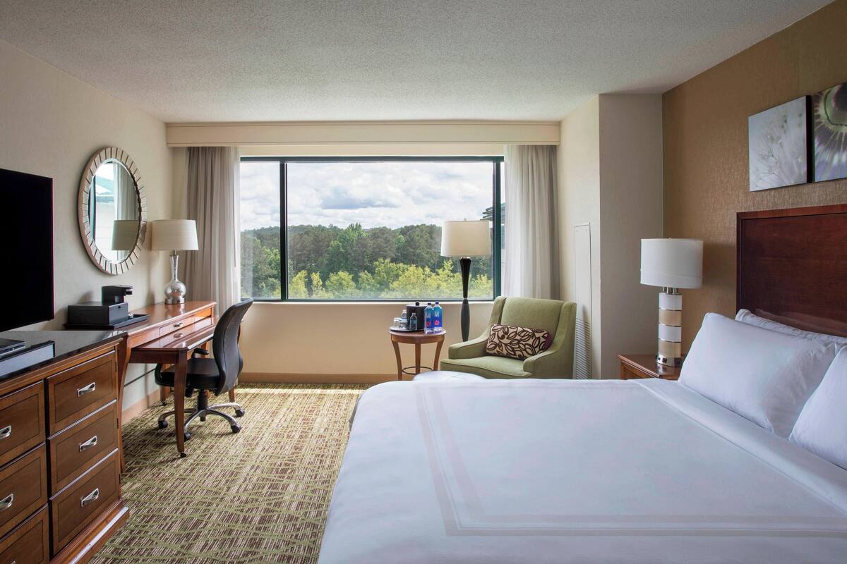 Habitación de hotel con una cama grande, un escritorio con computadora, una silla, un televisor y una ventana con vista a árboles.