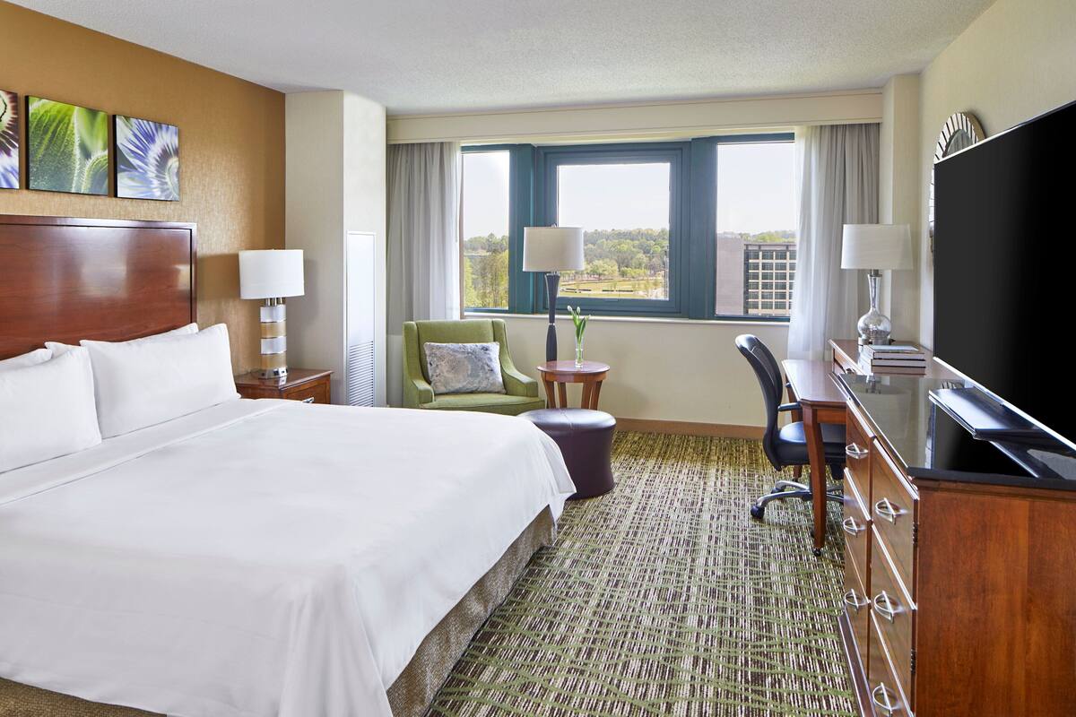 Habitación de hotel con una cama grande, un escritorio con computadora, un televisor y vistas a la ciudad.