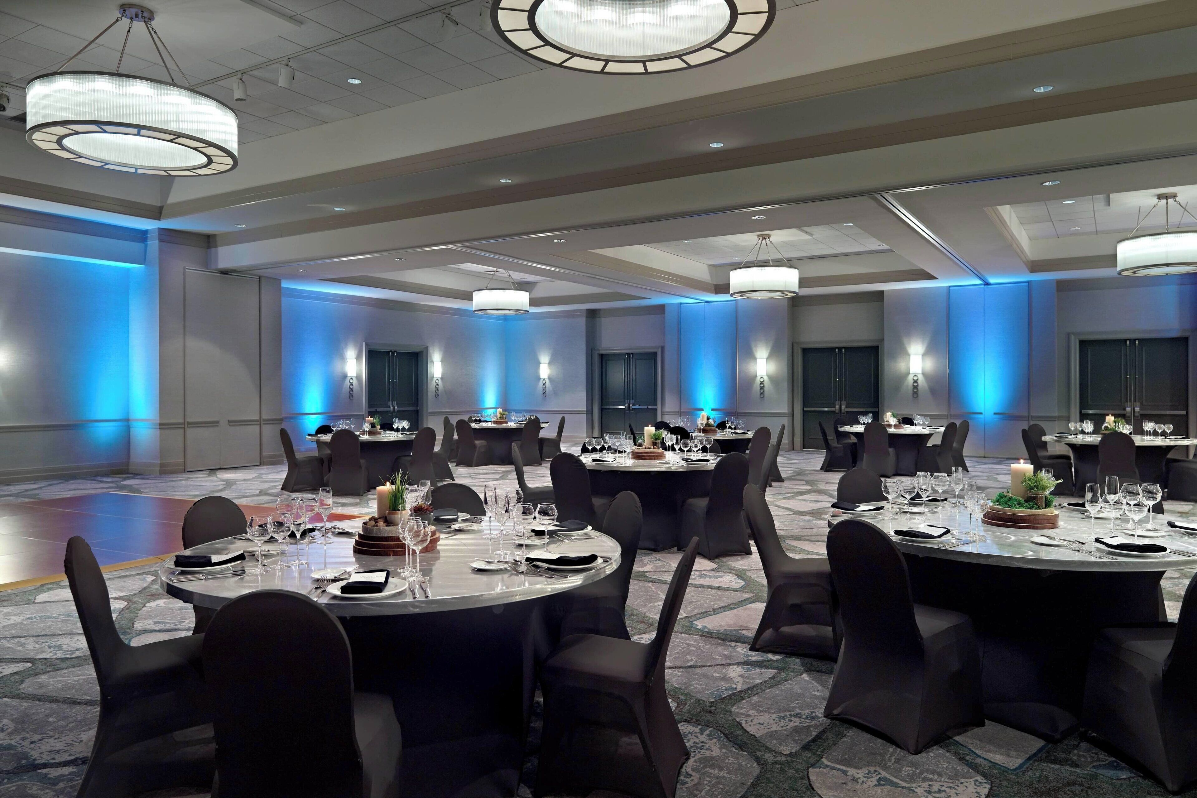 A spacious banquet hall with round tables, black chairs, and blue ambient lighting.
