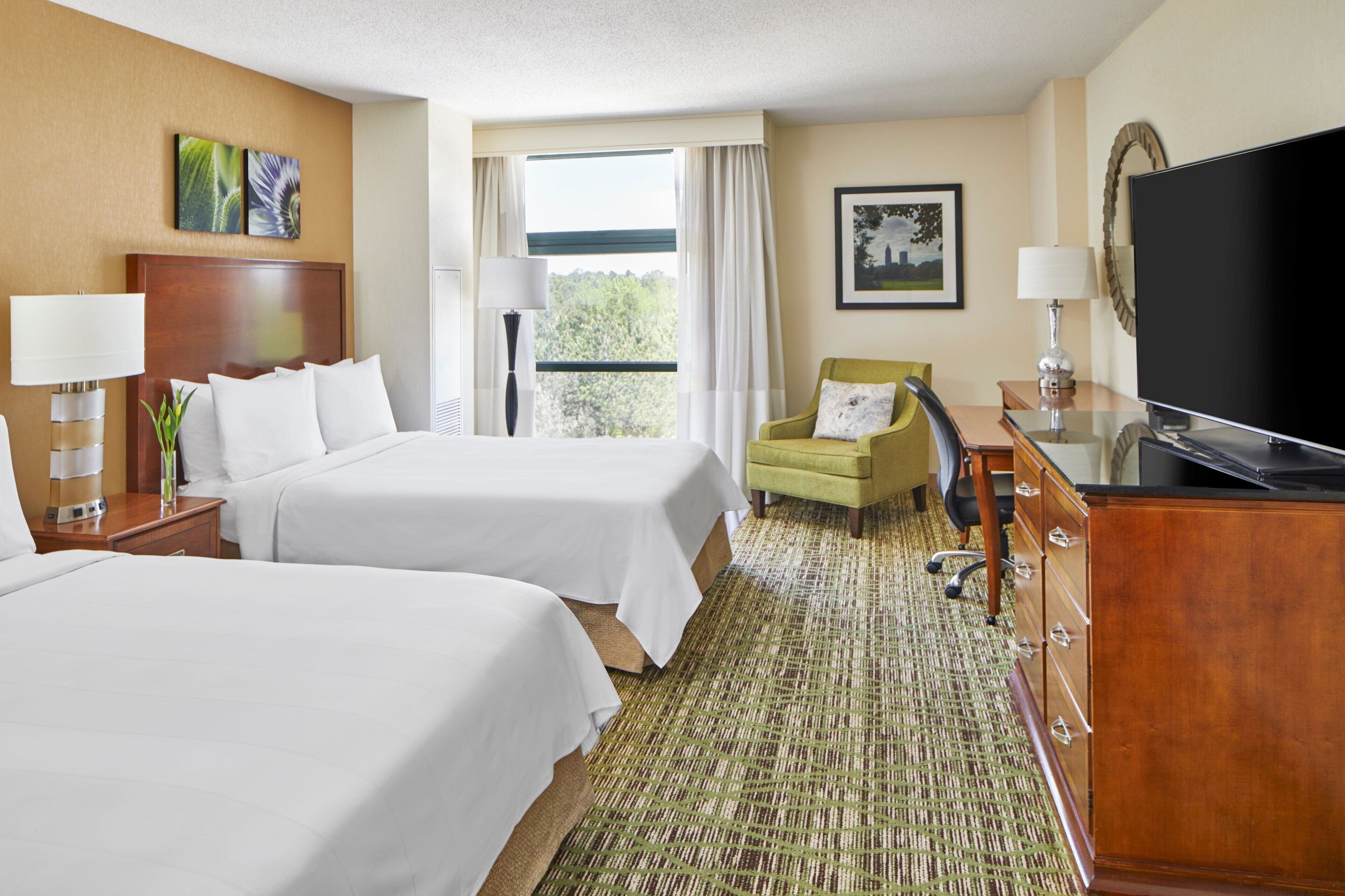 A hotel room with two beds, a television, a desk with a lamp, and a chair.