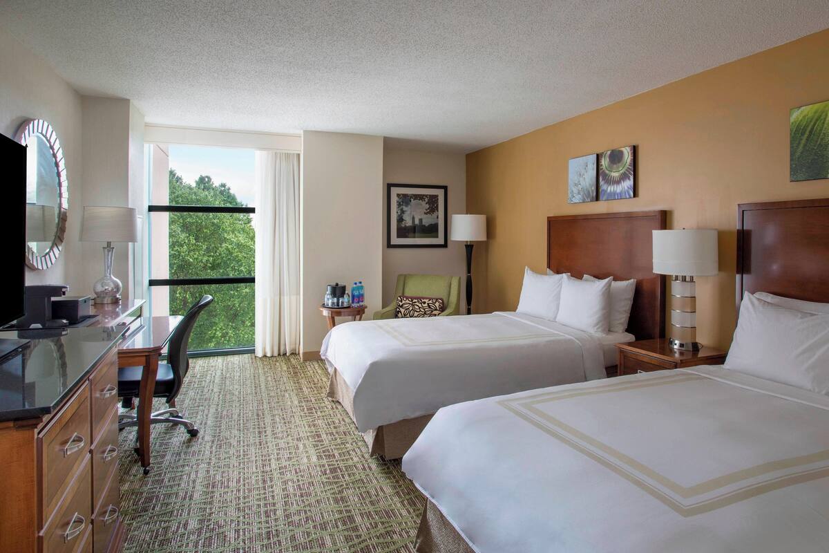 Habitación de hotel con dos camas, un escritorio con una computadora, una silla, una ventana con vista a árboles y un cuadro en la pared.