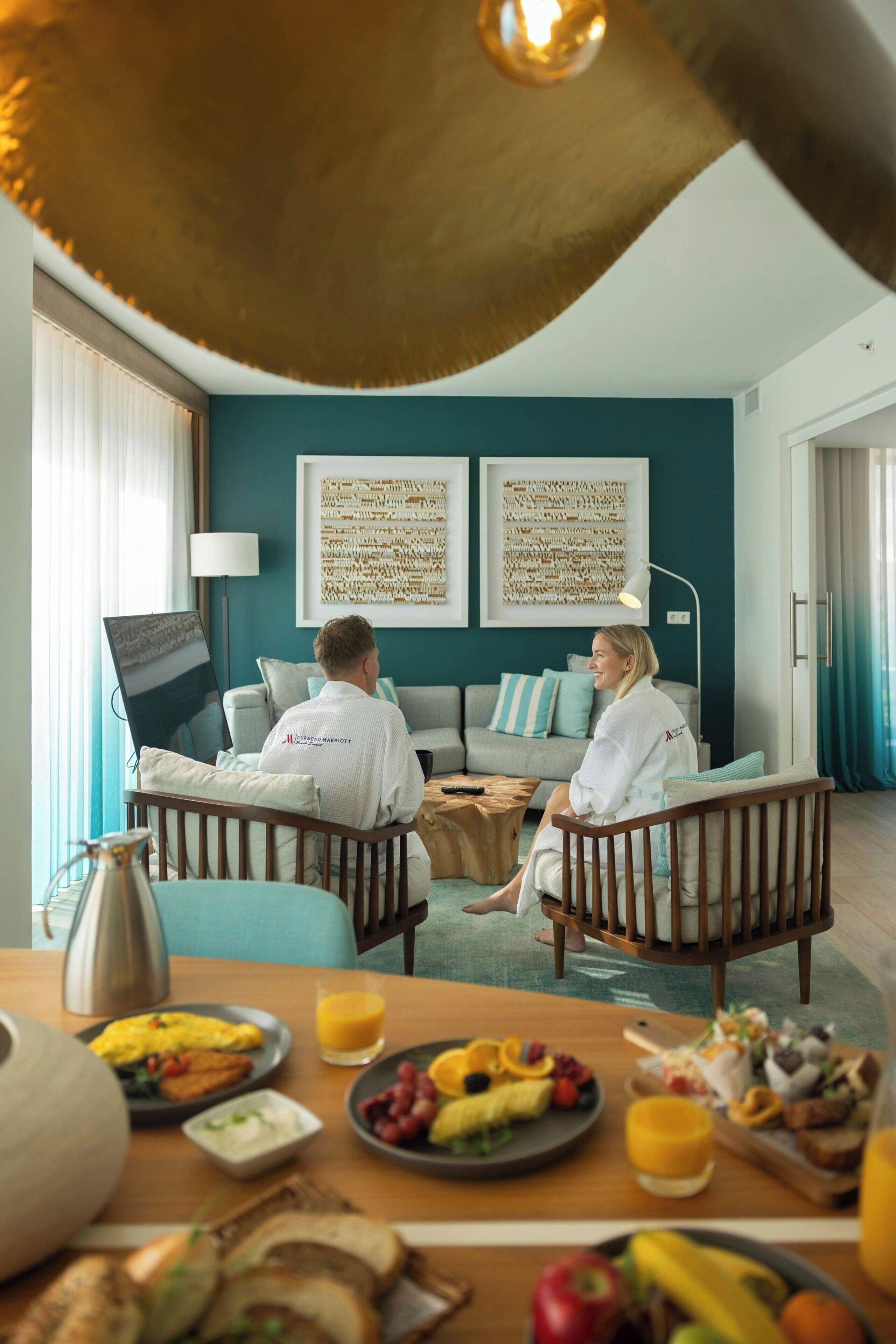 A hotel room with a dining area, two sofas, a coffee table, and a breakfast spread on the table.