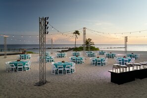 A beachside event setup with round tables and chairs, string lights, and a view of the ocean.