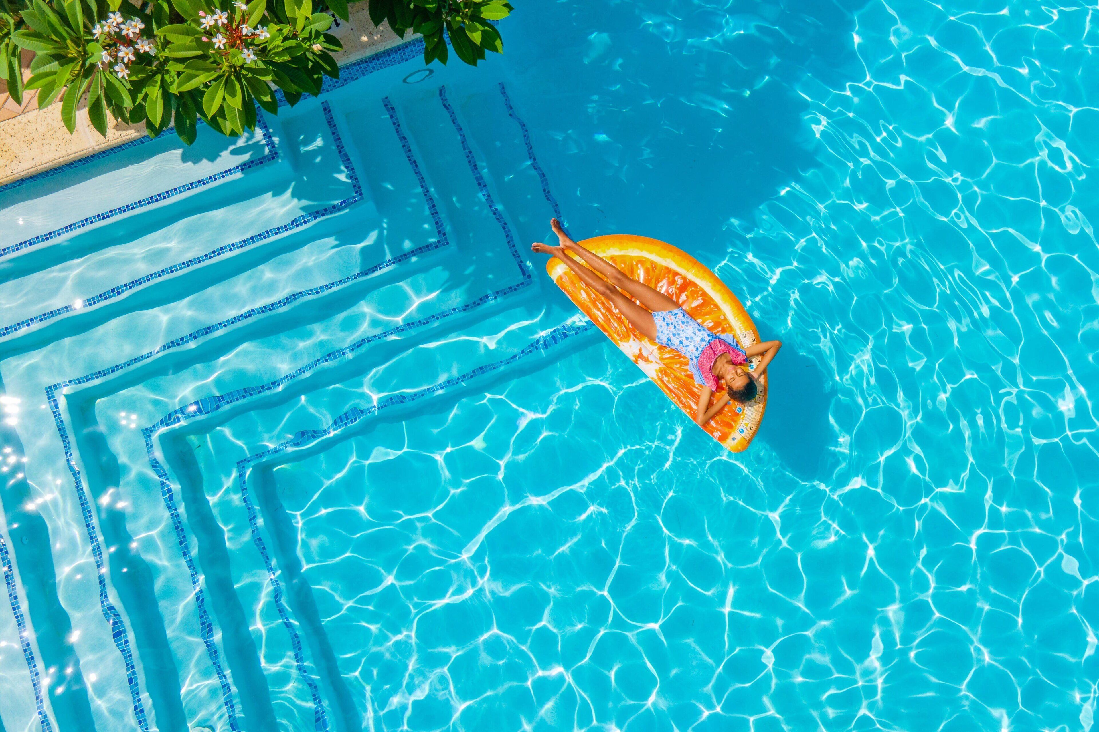 Eine Person treibt auf einem orangefarbenen Schwimmpfiff in einem klaren, blauen Pool mit gefliesten Stufen und grünem Grünwerk im Hintergrund.