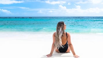 A person sitting on a white sandy beach with clear turquoise water and a blue sky in the background.