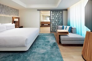 A hotel room with a large bed, a sofa, a TV, and a bathroom visible through a partition.