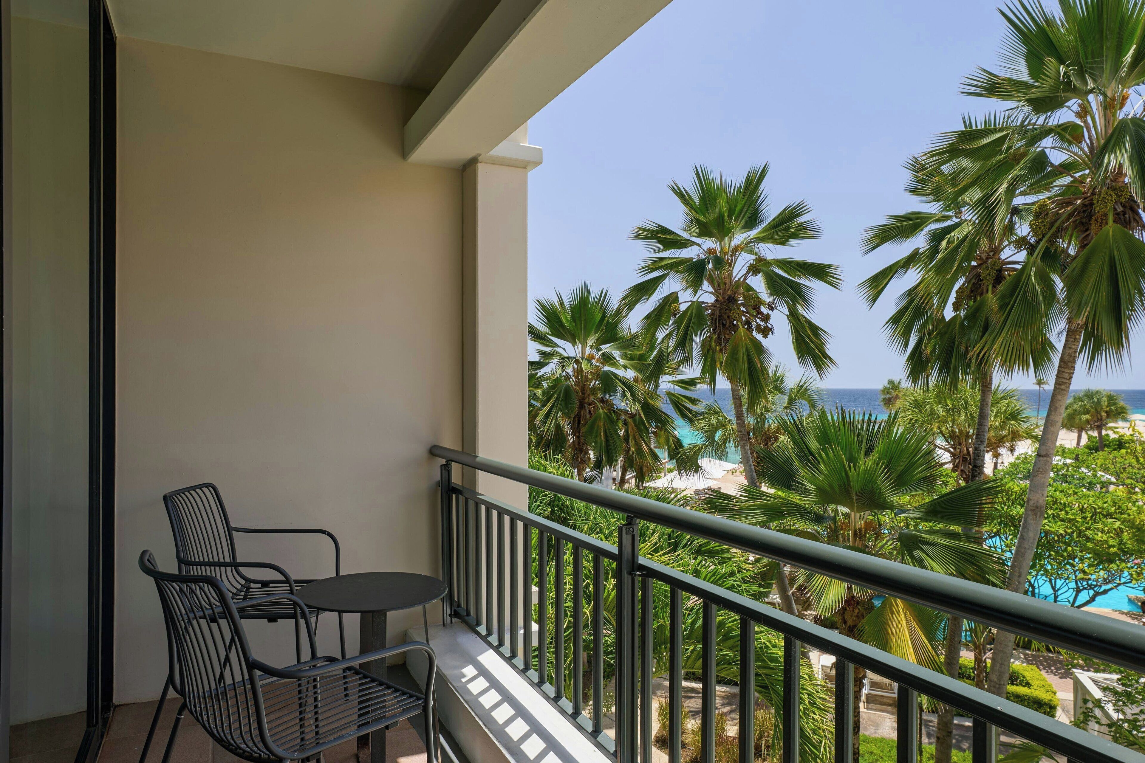 A balcony with a table and chairs, overlooking a beach with palm trees and a clear sky.