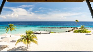 Blick auf einen Strand mit Holzterrasse, weißem Sand, klarem türkisblauem Wasser und Palmen.