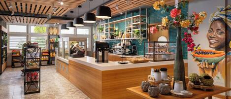 Interior moderno de una cafetería con mostrador de madera, estantes con aperitivos, una máquina de café y un mural de una mujer sonriendo que sostiene un tazón de fruta.