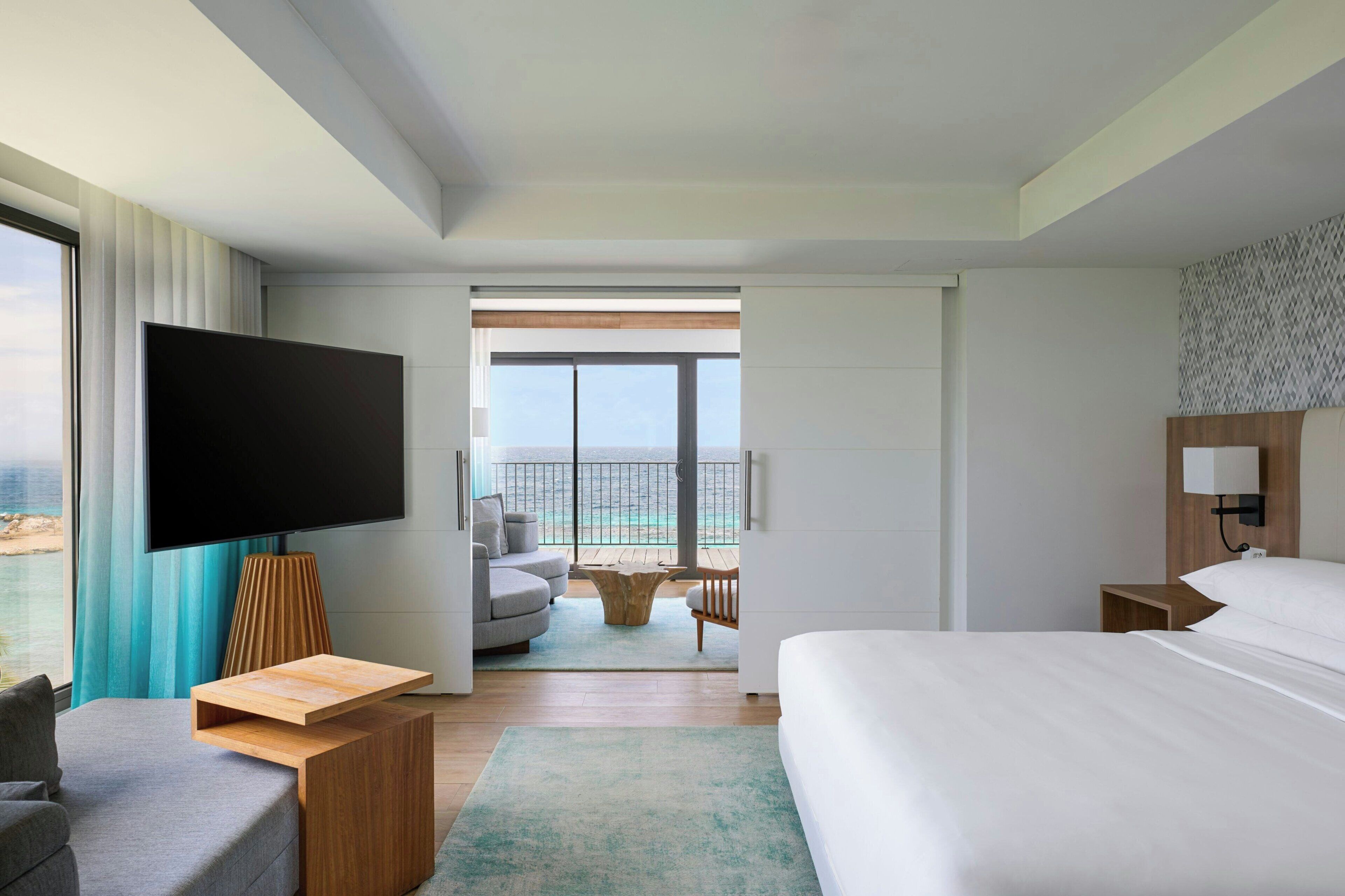 Una habitación de hotel moderna con una cama grande, un televisor de pantalla plana, un área de descanso con una mesa redonda y un balcón con vistas al mar.
