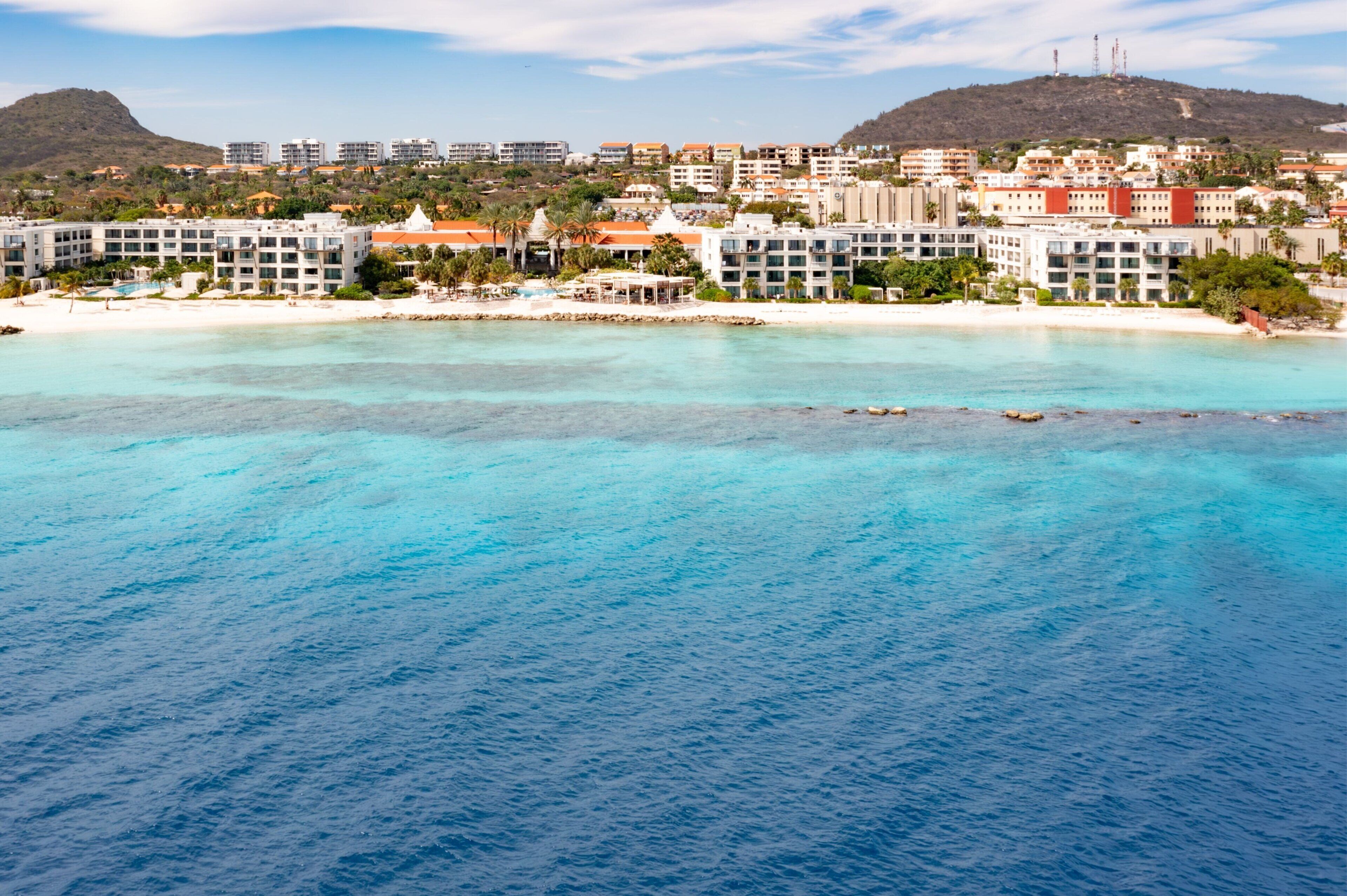 Un hotel frente al mar con varias edificaciones, una playa y aguas azules cristalinas.