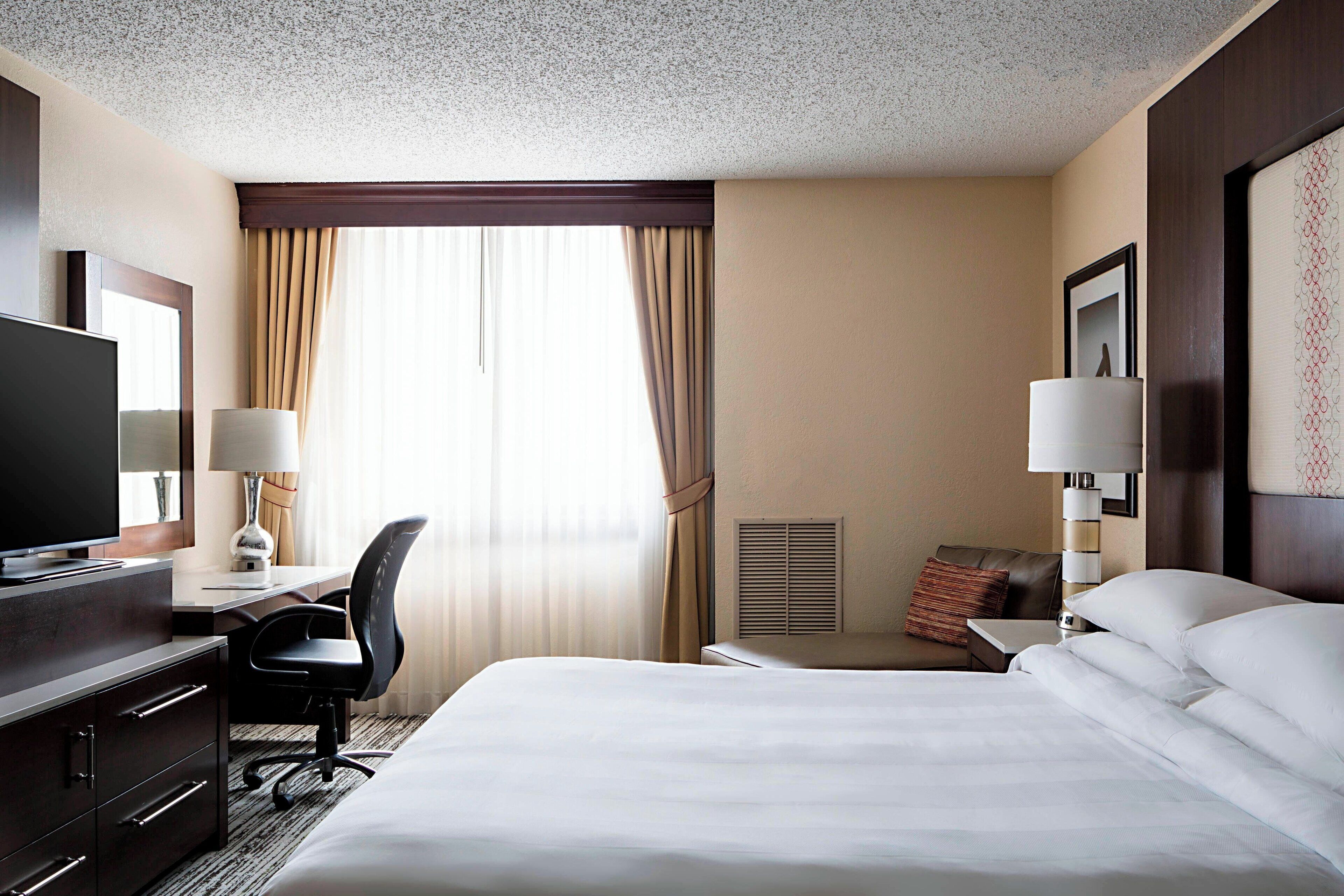 A hotel room with a large bed, a desk with a chair, a television, and a window with curtains.