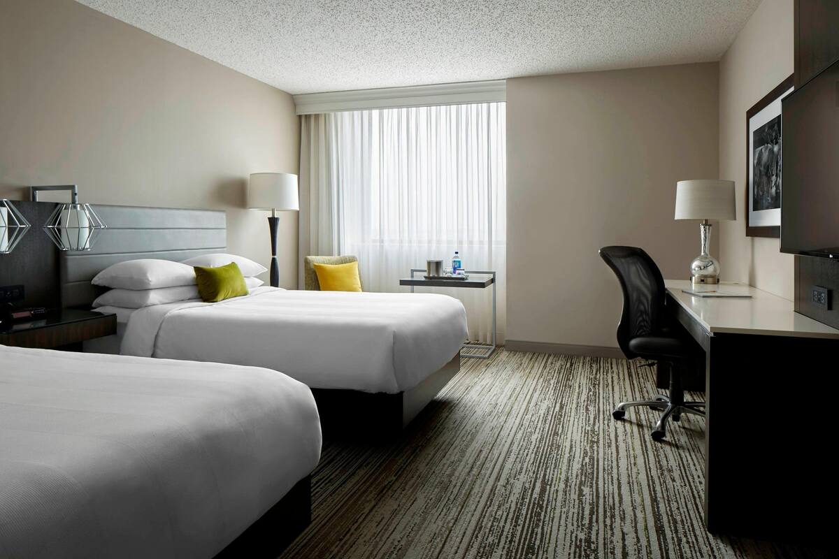 A hotel room with two beds, a desk, a chair, a TV, and a window with curtains.