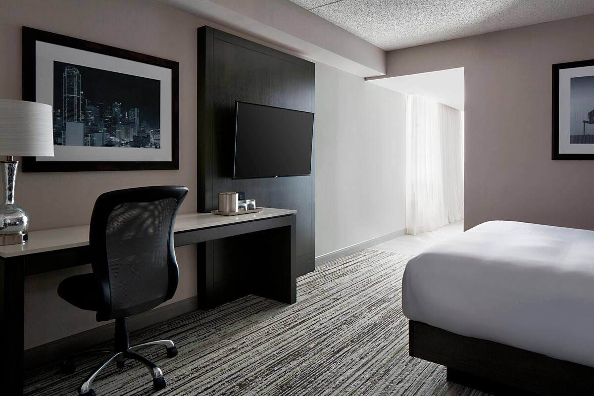 A hotel room with a bed, a desk with a chair, a flat-screen TV, and a framed cityscape picture on the wall.
