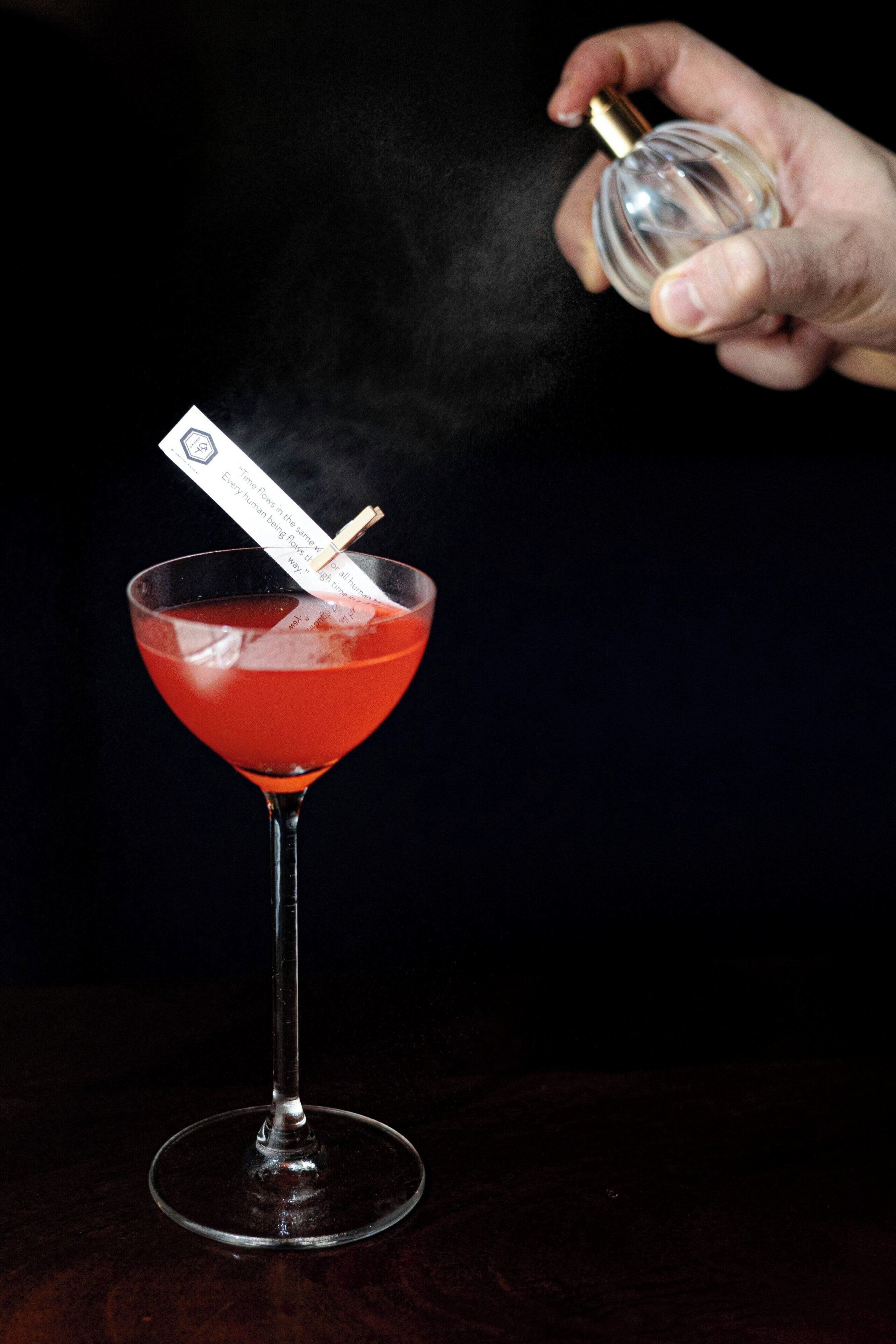 A hand holding a bottle of perfume, spraying it onto a cocktail in a martini glass.