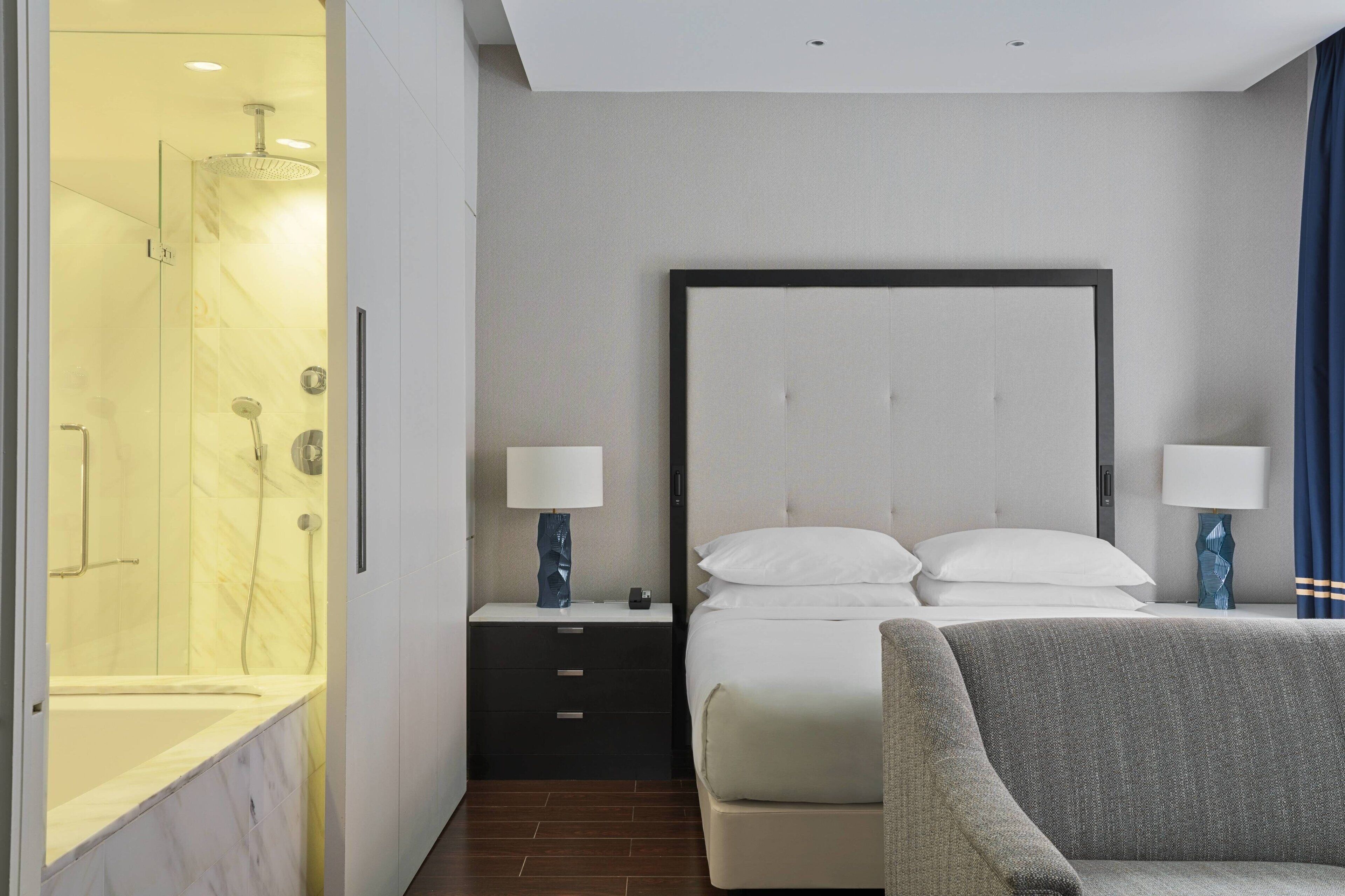 Een moderne hotelkamer met een marmeren badkamer, een groot bed met witte lakens, twee nachtkastjes met lampen en een grijze bank.