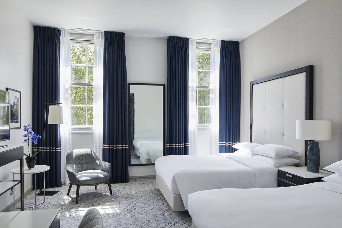 A hotel room with two beds, a desk, a chair, and large windows with curtains.