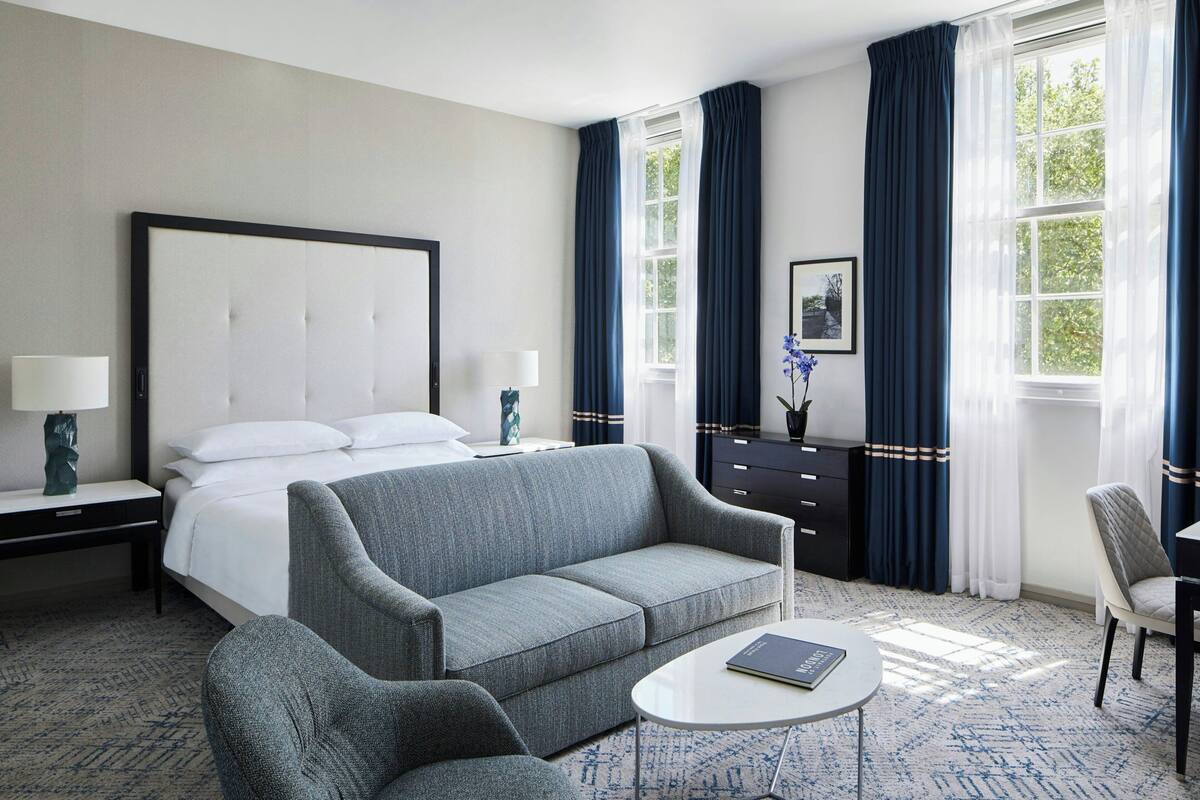 A hotel room with a bed, a grey sofa, a white coffee table, a desk with a lamp, and a chair.