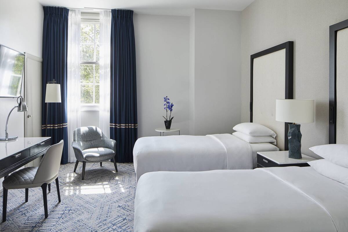 A hotel room with two beds, a desk, a chair, a lamp, a small table with a vase of flowers, and a large mirror on the wall.
