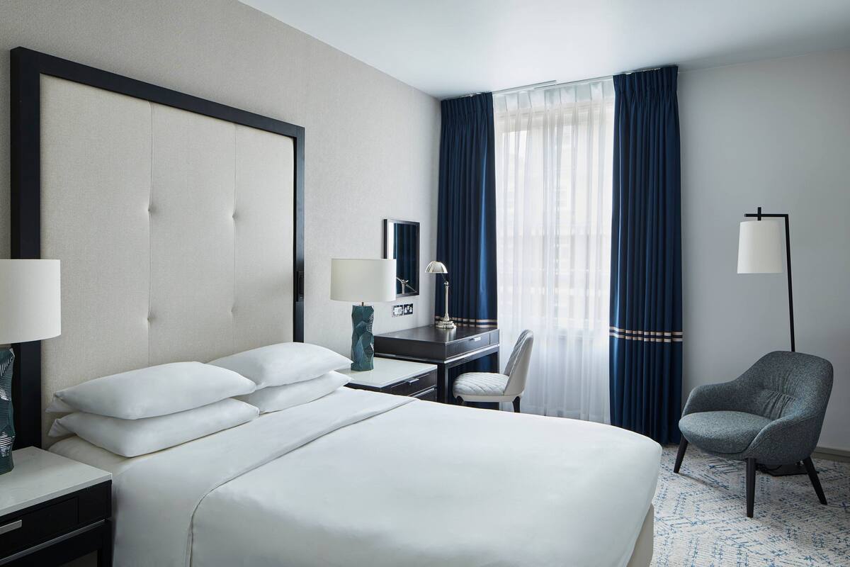 A hotel room with a large bed, a desk with a chair, a lamp, and a view of the outdoors through a window with curtains.
