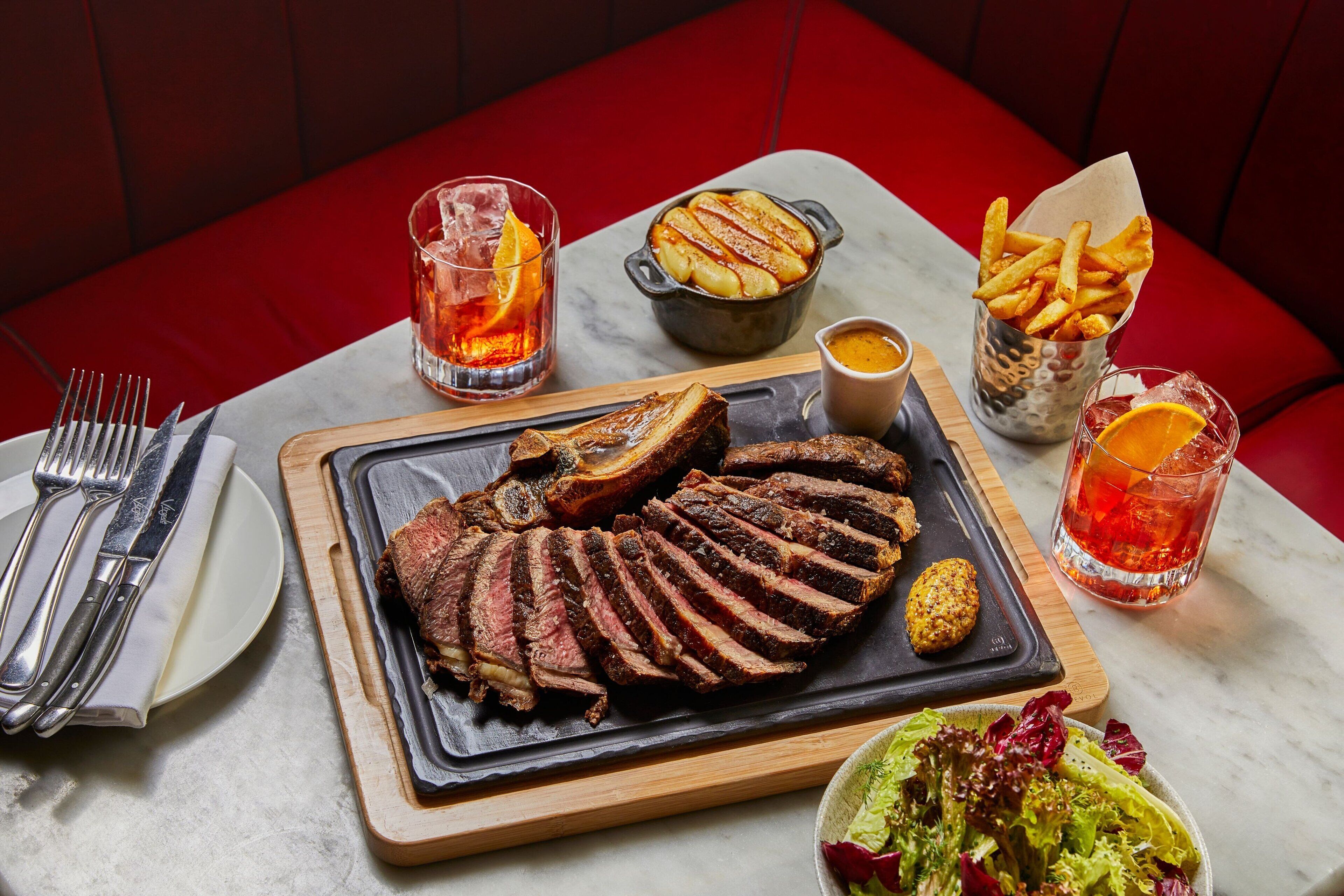 Una mesa de comedor con un bistec, papas fritas y ensalada, acompañada de bebidas y cubiertos.